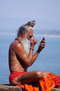 A Sadhu sits by a river and applies his tilaka (forehead marking) while using his mobile phone as a mirror.