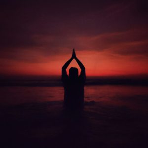 Silhouette of a man at sunset with arms raised above his head in prayer position.